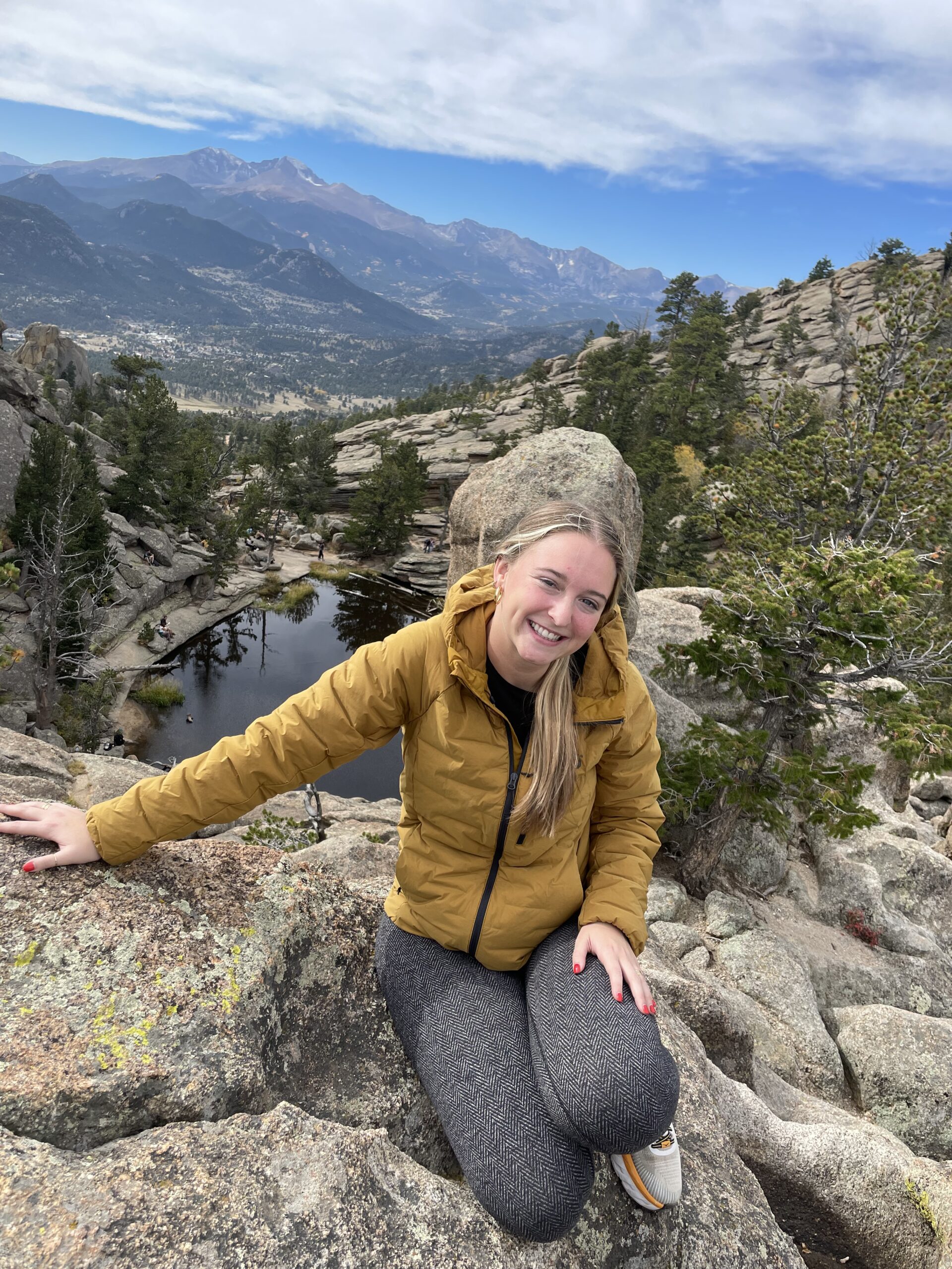 Ms. Beach sitting on a rock with a yellow jacket on Infront of a mountain landscape.