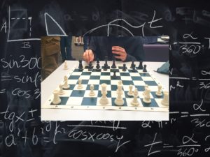 A chess board, mid-game, is pictured against a chalkboard with mathematical calculations drawn on it