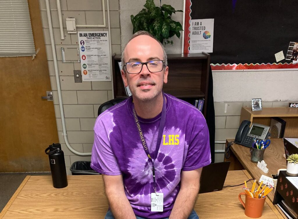 Mr. Fisher poses, sitting on his desk